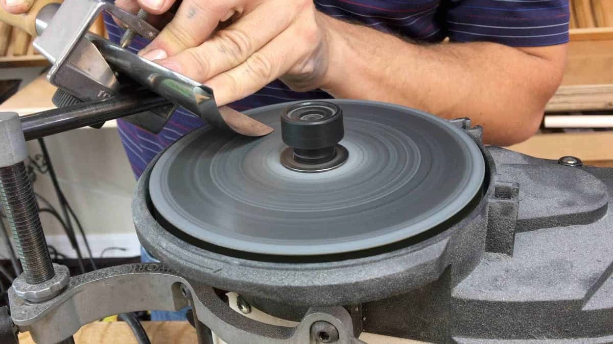 Metal jig holds the big gouge to the grey wheel while a hand steadies the blade