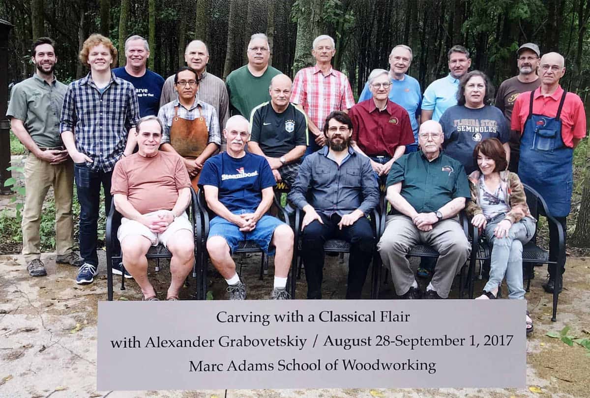 Class group photo with a printed sign reading Carving with a Classical Flair with Alexander Grabovetskiy, August 2017