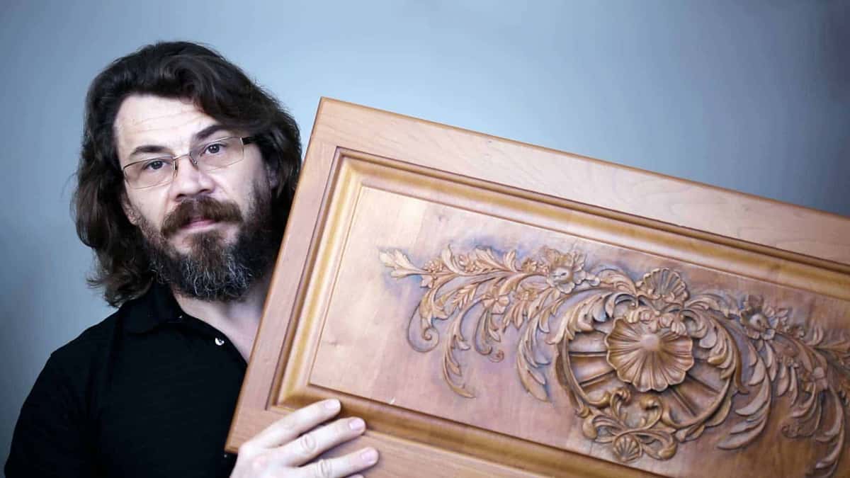 A bearded man holds up the cherry door panel, deep shell at the middle, acanthus trailing off it