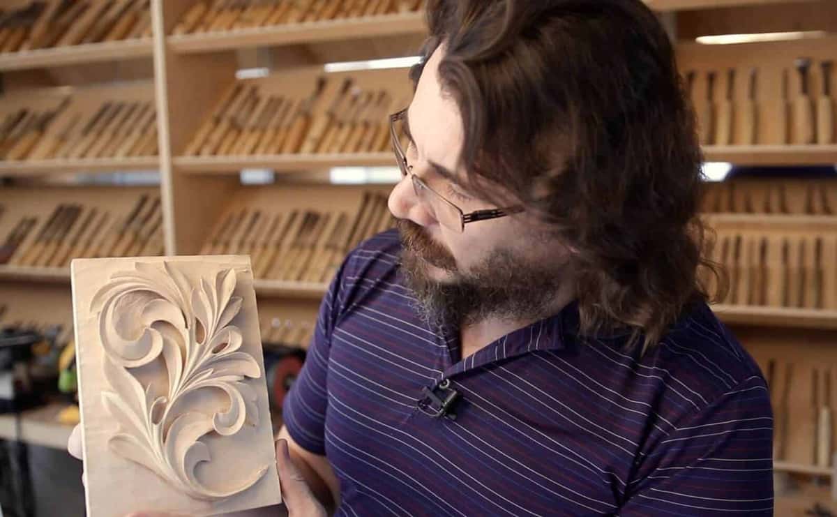 A pale board on a ball vise carries a deep acanthus scroll; the board's square edge and full thickness are in view.