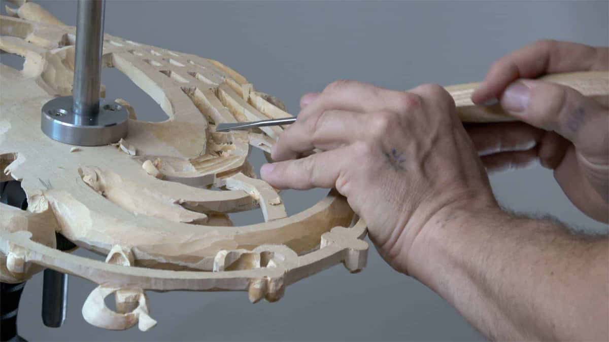 A large pierced rosette in pale wood on a vise post, its openings carved right through, a skew chisel at one edge.