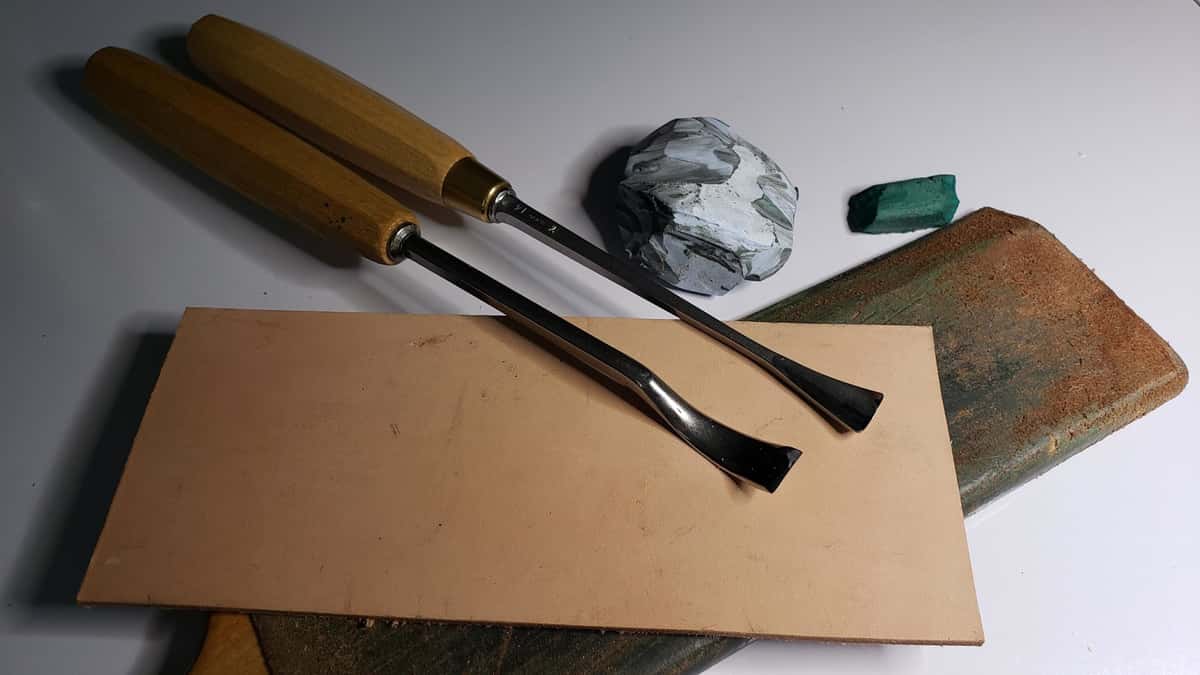 Two bent gouges lying on a bare leather strop beside a bar of green honing compound and a loaded strop block