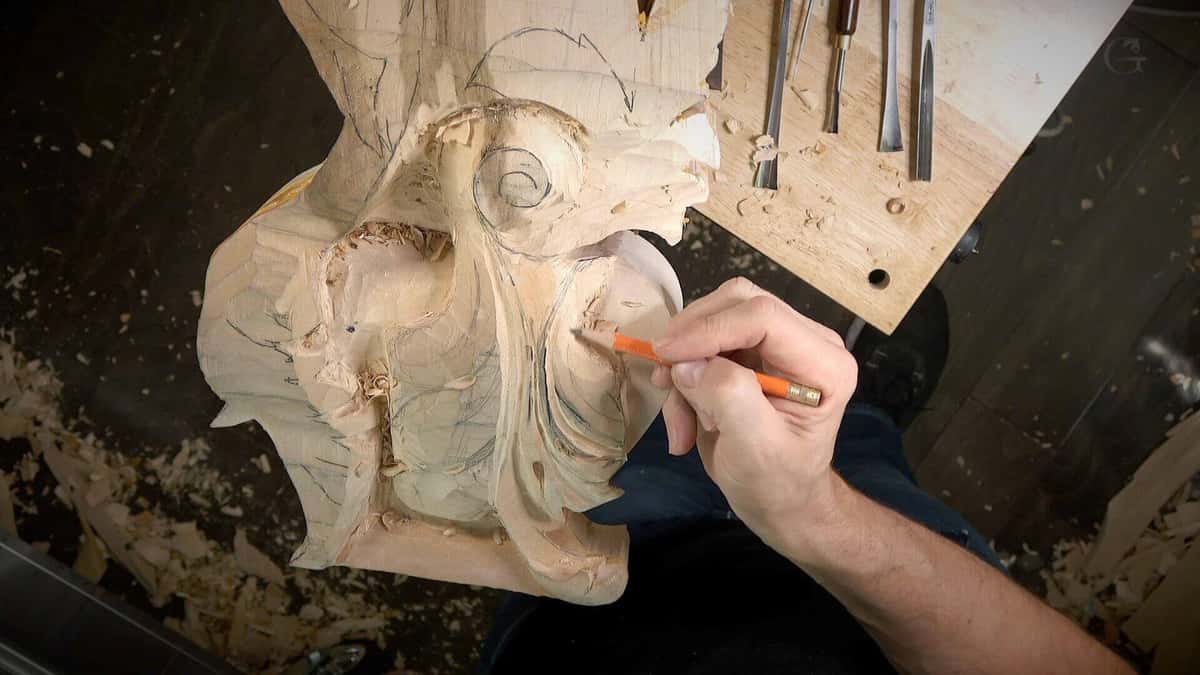 An orange pencil redraws a line on a pale carved scroll blank, five gouges laid out on the board.