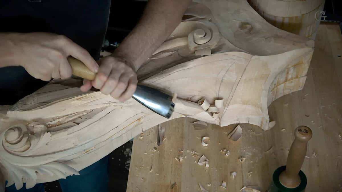 A wide firmer gouge shapes the eye of a scroll while the mallet waits