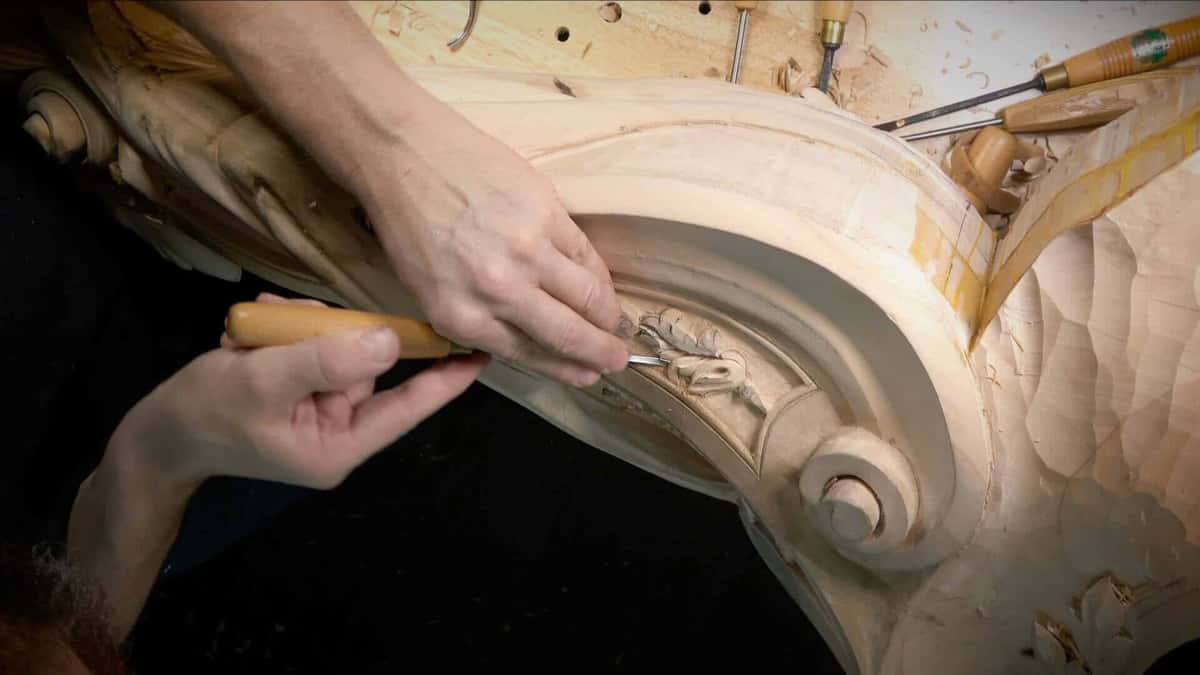 Two hands work a small carved band on a curved rail, with gouges racked on the bench behind.