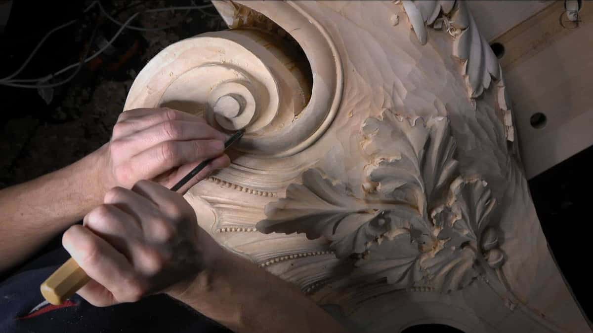 Hands carve the spiral eye of a large volute, with a bead row and deep undercut acanthus beside it.