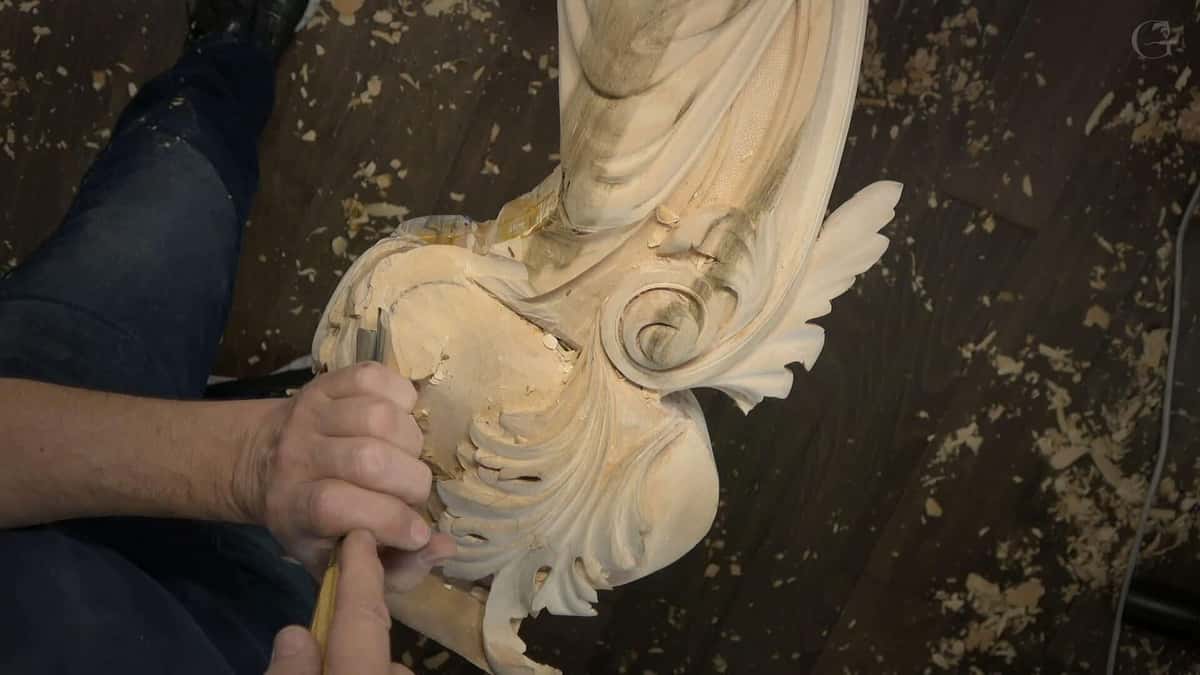 A pale carved scroll and leaf panel lying face up on a dark plank floor, chisel in hand, chips scattered.