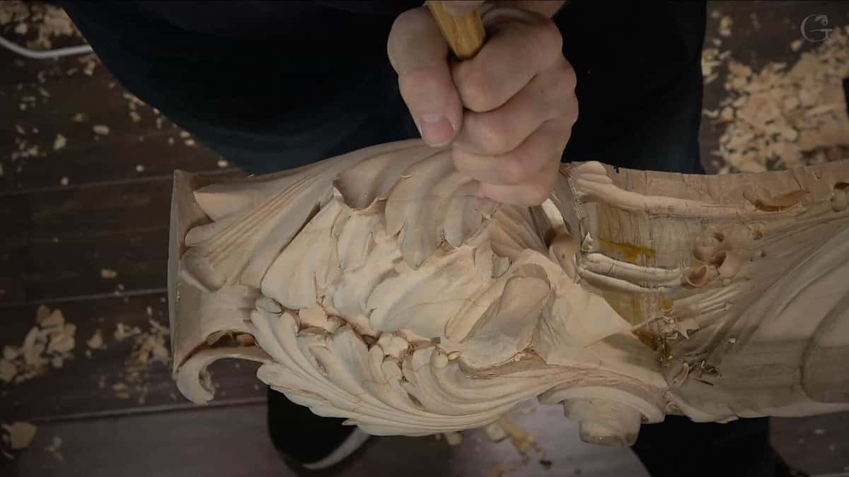 A long carved leaf held up mid-work, still showing broad tool facets, with chips over the floor below.