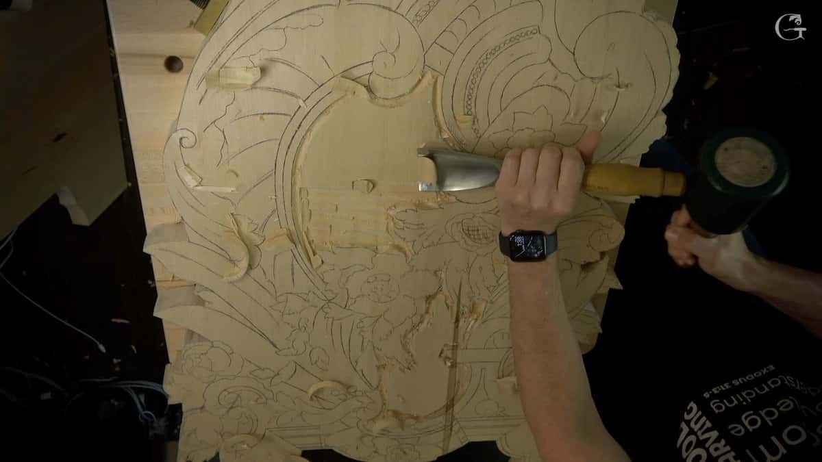A wide gouge driven by a green mallet takes a pass across a big pale panel drawn all over in pencil.