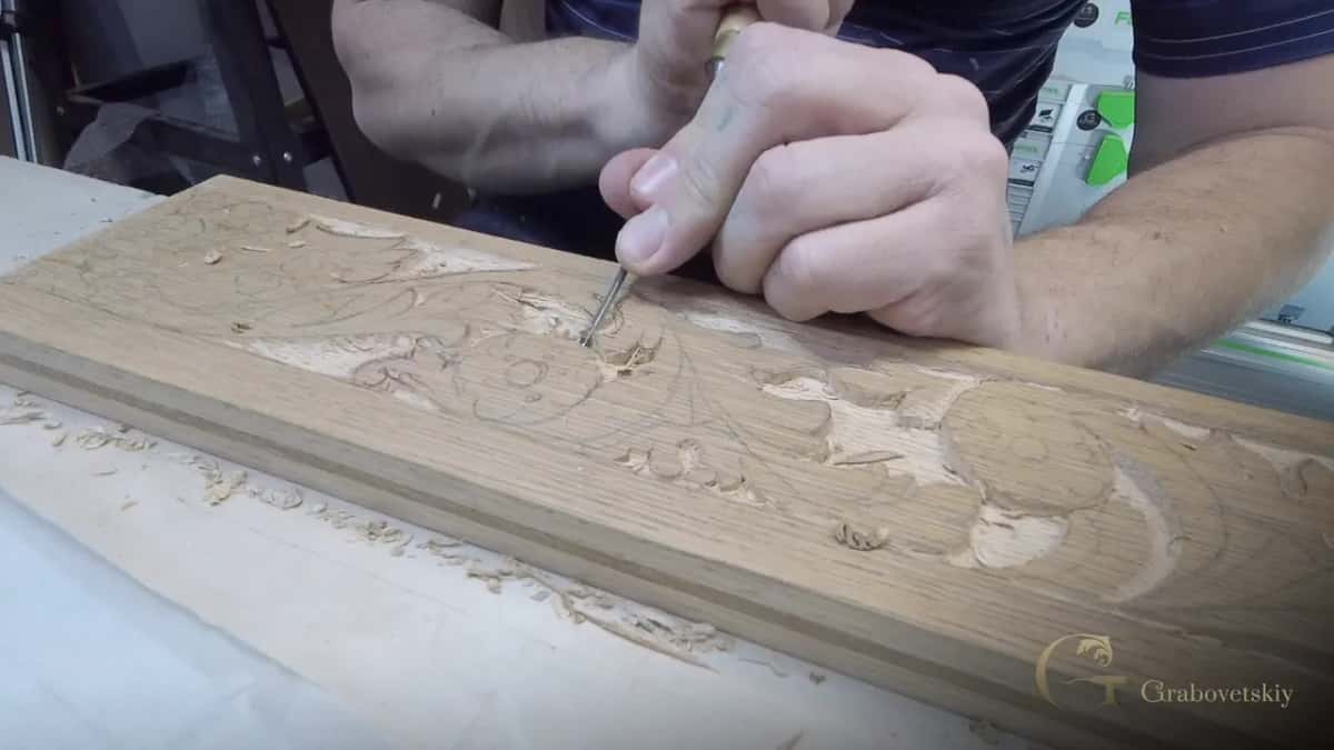 Both hands wrapped around one small chisel, pushing into the scroll of an oak apron with pencil lines still showing.