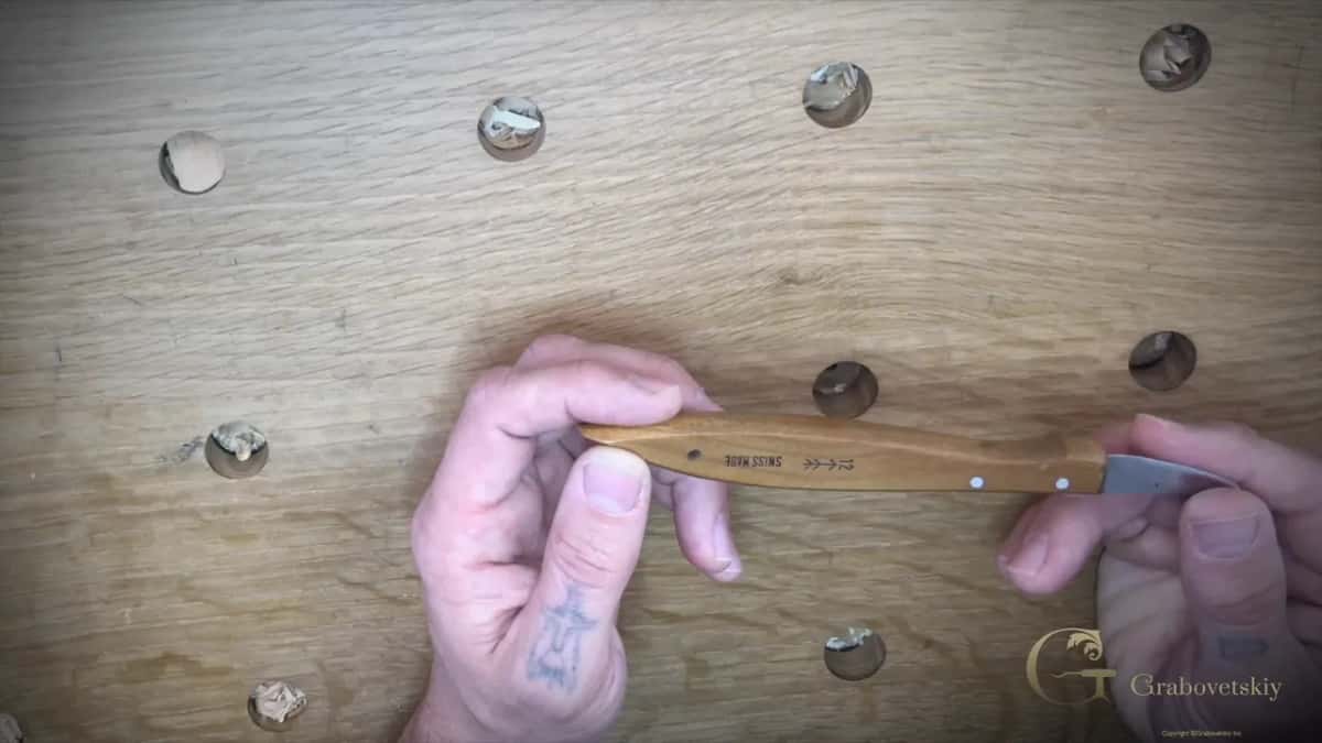 Two hands hold a Swiss-made No. 12 chip carving knife over an oak bench dotted with dog holes and chips.