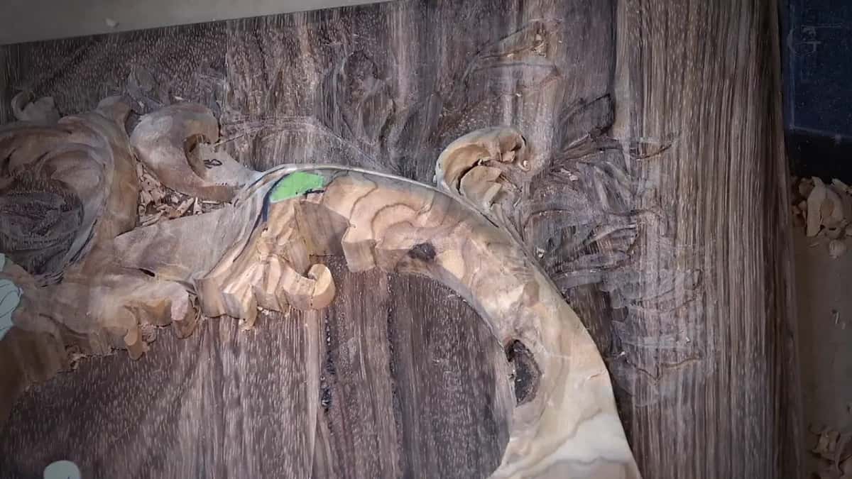 A pale carved scroll and leaves stand out of a dark hardwood panel, wood chips piled at the left edge.