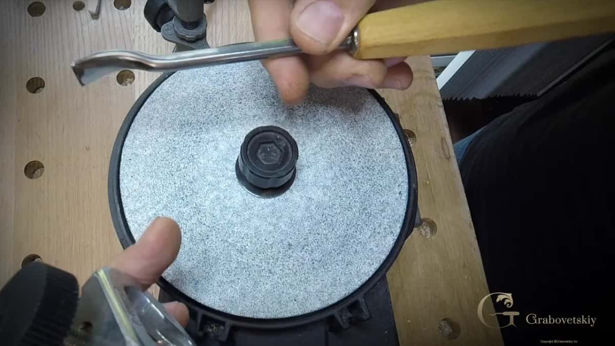 Two hands hold a bent gouge flat against a grey honing disc clamped at the bench.