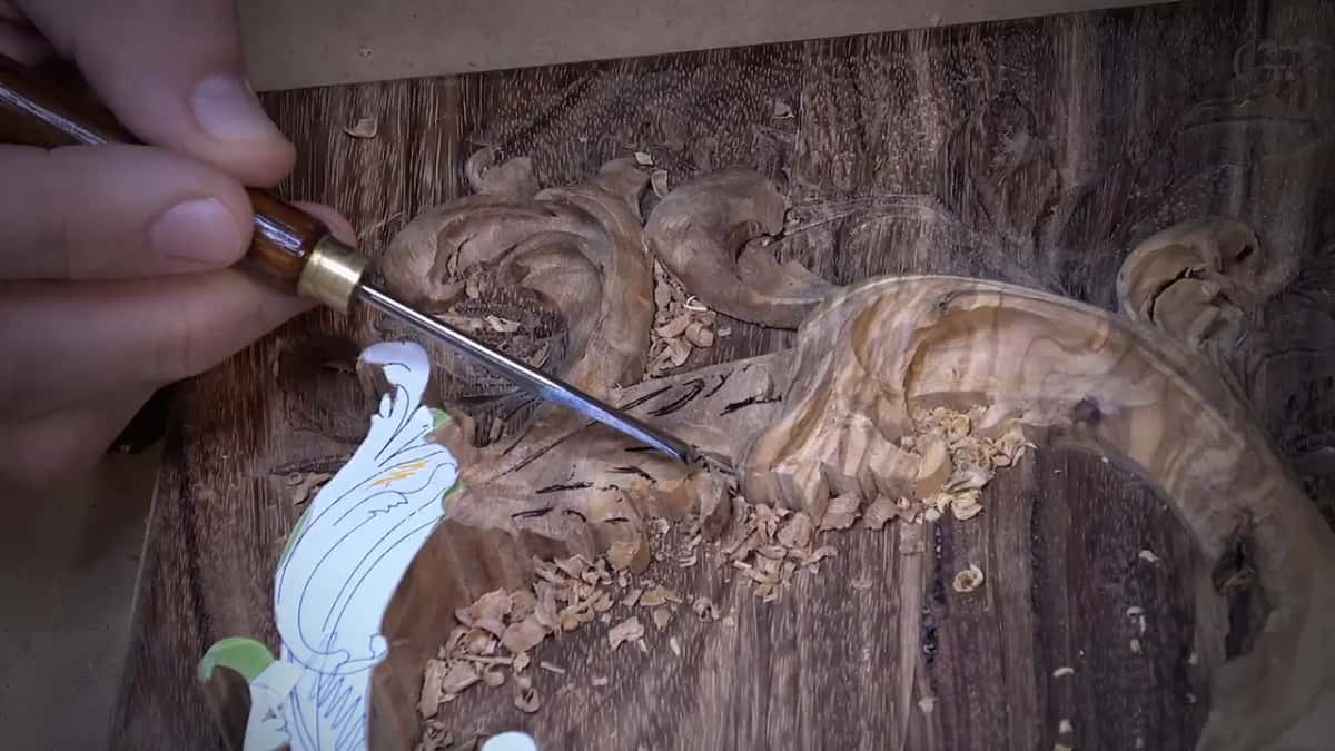A long thin gouge carving a figured scroll on a dark panel, shavings piled around the work.