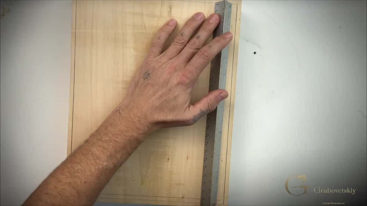 A hand holds a steel rule against a pale flat board marked with a pencil border for a carved panel.