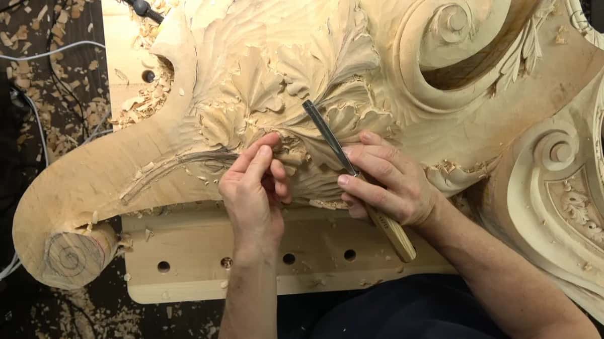 Hands set a chisel into a large pale volute; the sawn end of the blank carries a pencilled spiral.