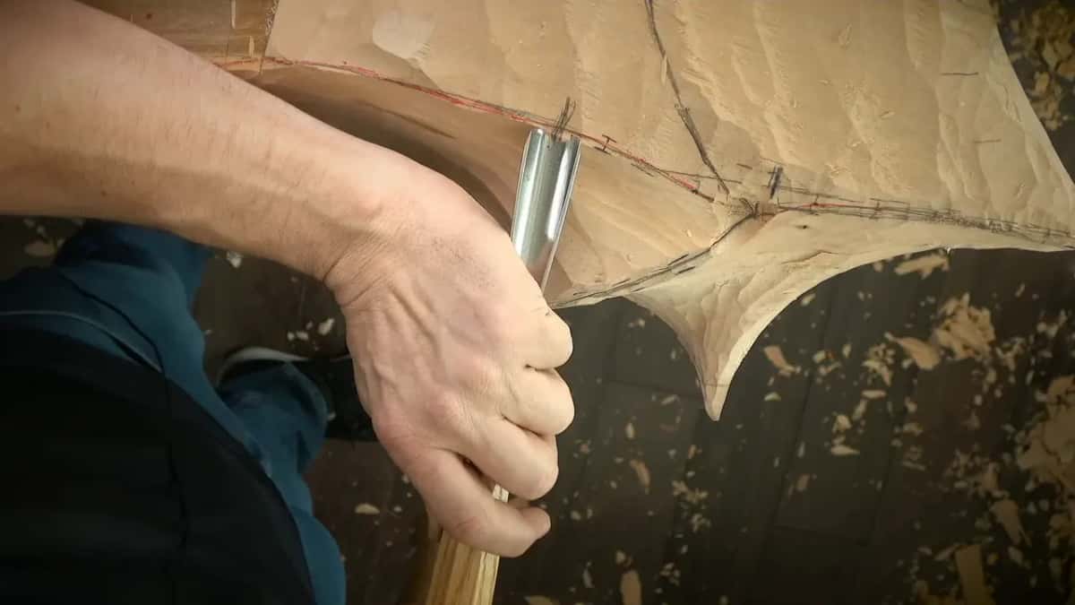 A fist grips a long gouge, its bright bevel planted in a pale blank marked out in red and pencil.