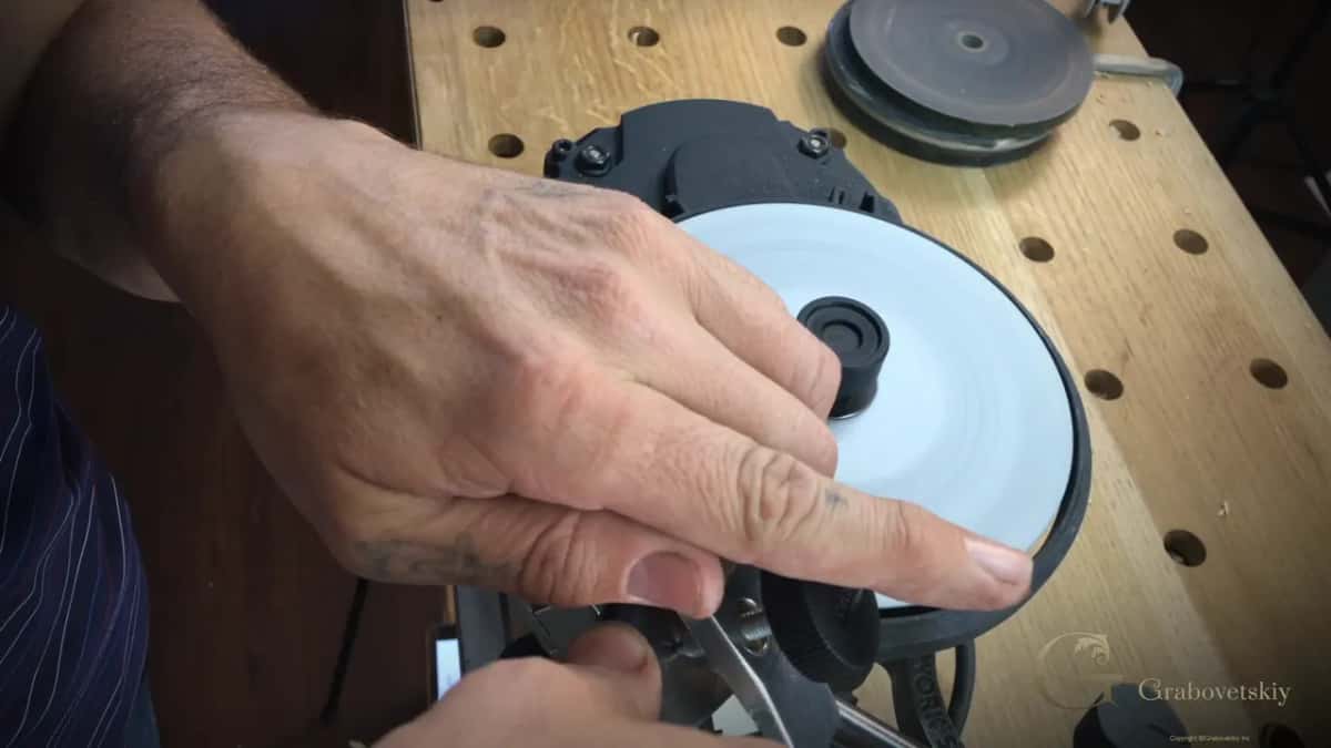 A hand steadies the white honing wheel of a sharpening machine, a gouge waiting in the jig below it.