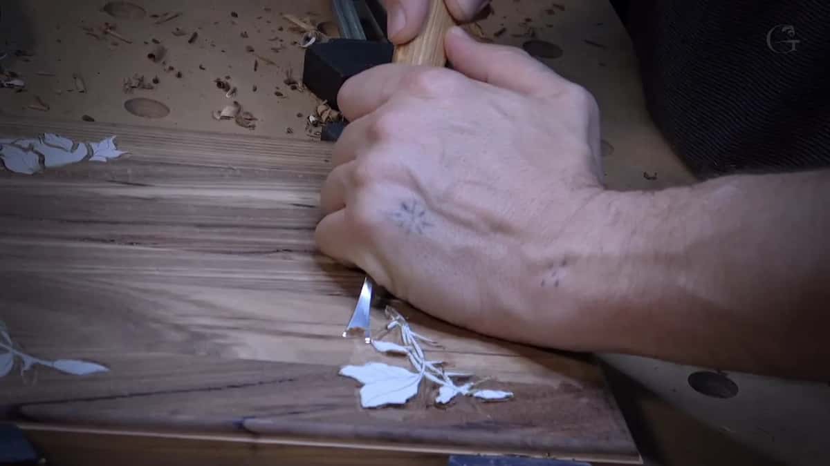 A mallet raised over a gouge tracing a leaf outline into a dark figured box lid, the design painted white.
