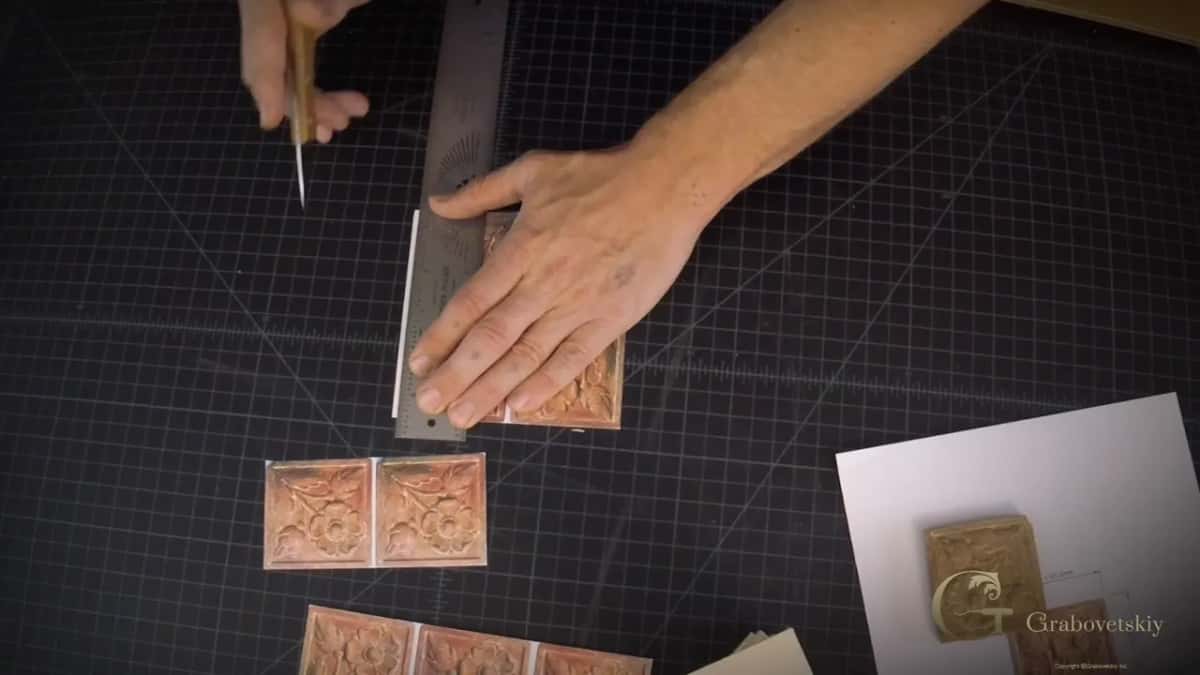 Printed tiles of a flower panel spread on a dark work mat, a steel rule pinned down and a small blade waiting.
