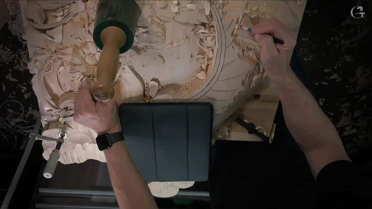 A mallet in one hand and a pen in the other, with a tablet propped upright on the bench as the reference.
