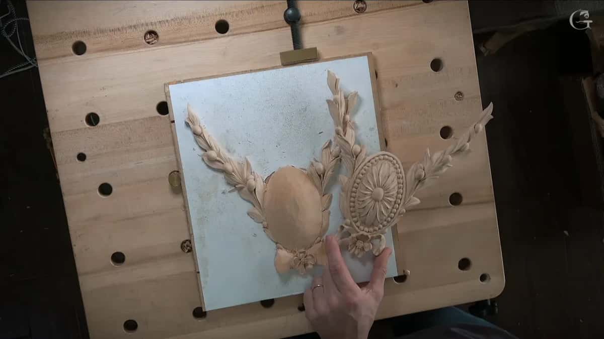 A hand slides a carved rosette next to a laurel spray on the board, trying the two pieces together.