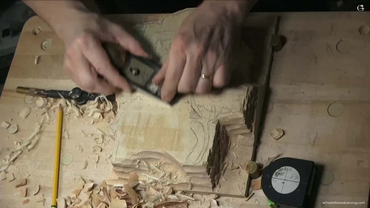 Two hands push a small metal plane across a board on the bench, with fresh shavings piled around it.