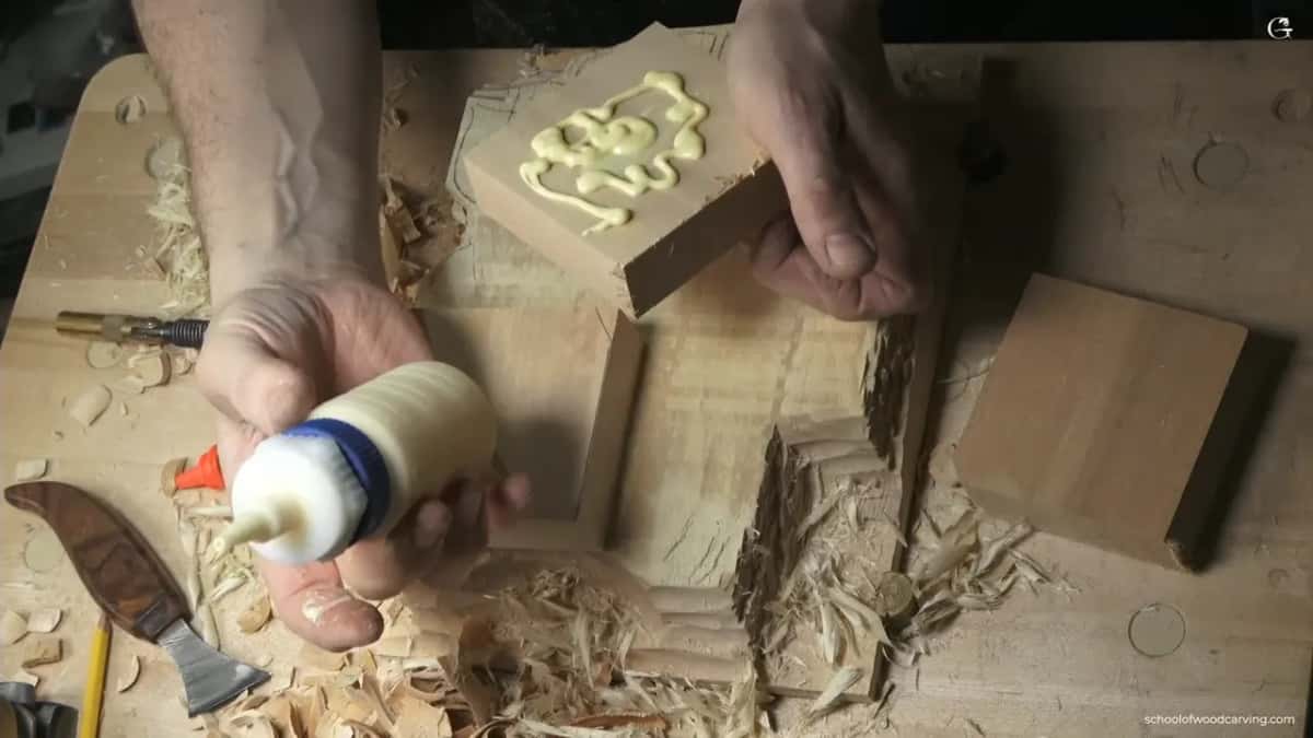 One hand holds a glue covered block, the other a glue bottle, while a plain block waits on the bench.