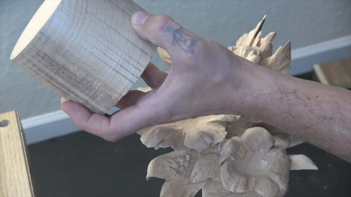 A hand lifts a round blank beside a group of carved flowers so the two can be compared.