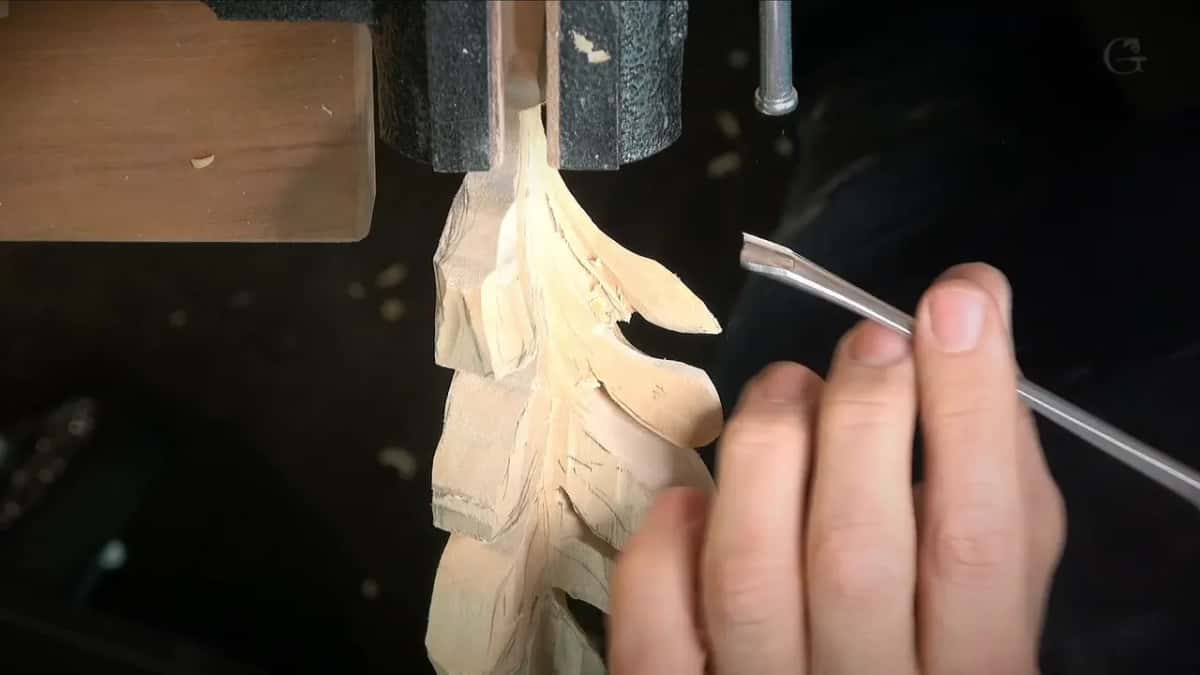 The tip of a small curved blade comes in toward a slim carved leaf clamped upright in the vise.