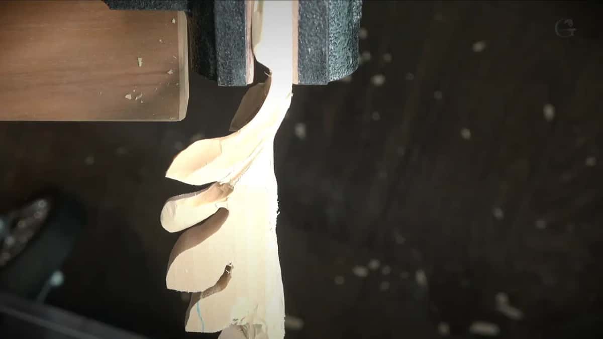 A carved leaf held in a padded vise is thin enough that light rings its edge against the dark shop.