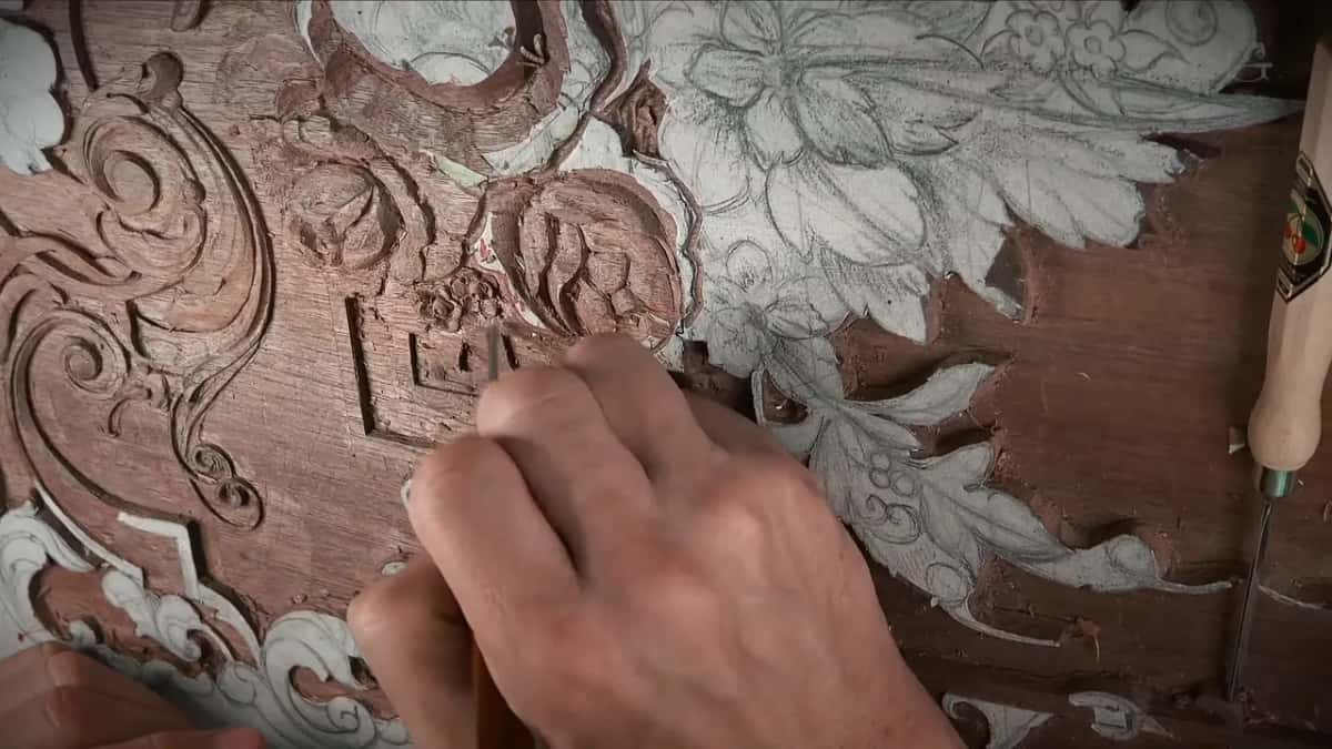 Right beside a pencil drawing still stuck to the dark panel, a hand carves the same pattern in wood.