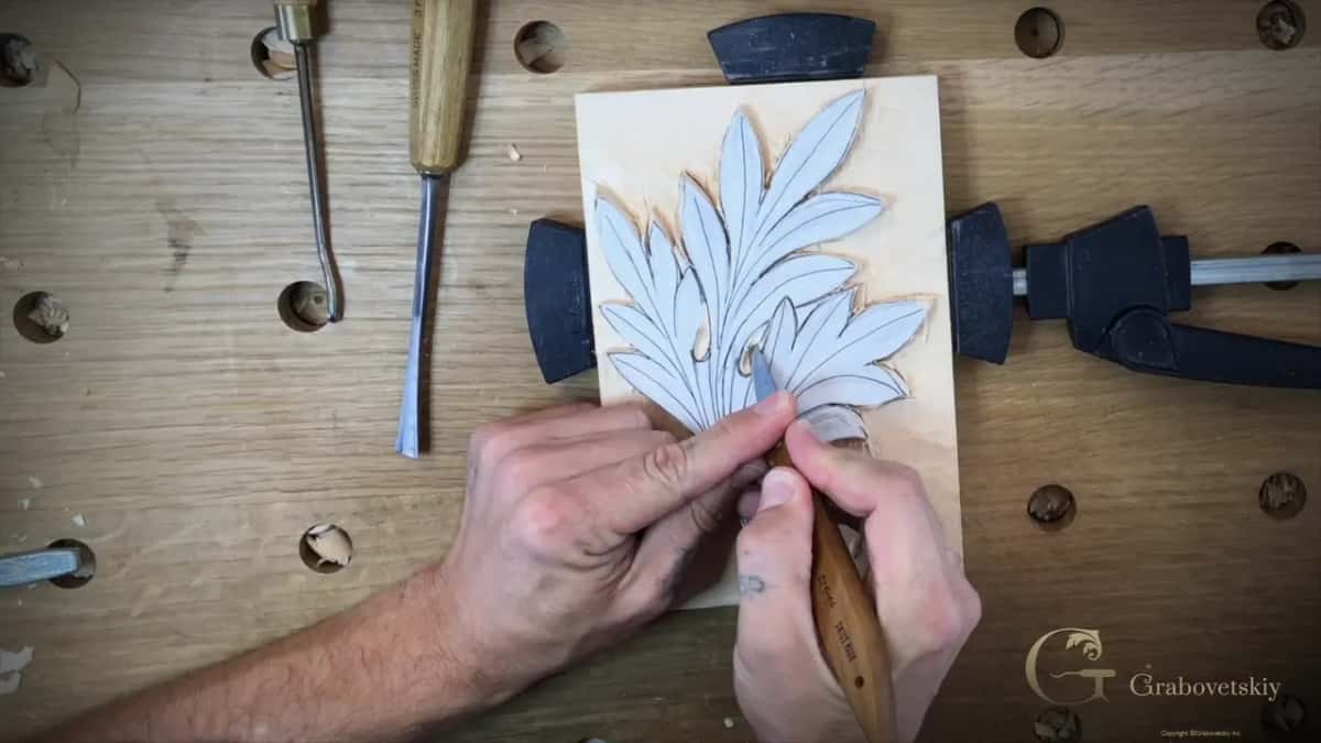 A knife held in both hands works the tip of a leaf pattern glued to a pale board between two clamps.