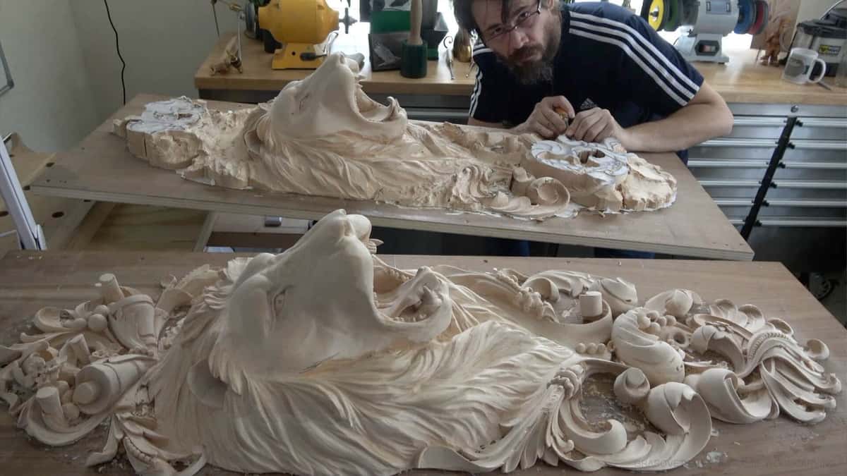 Two long lion head panels in pale wood lie on shop benches, one finished and one still being roughed out.