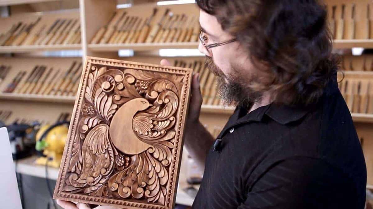 A person admires a beautifully carved wooden piece featuring intricate designs of a bird and floral patterns. In the background, you can see lots of carving tools lined up neatly. This showcases the art of wood carving and highlights the enjoyment found in taking wood carving classes and lessons.