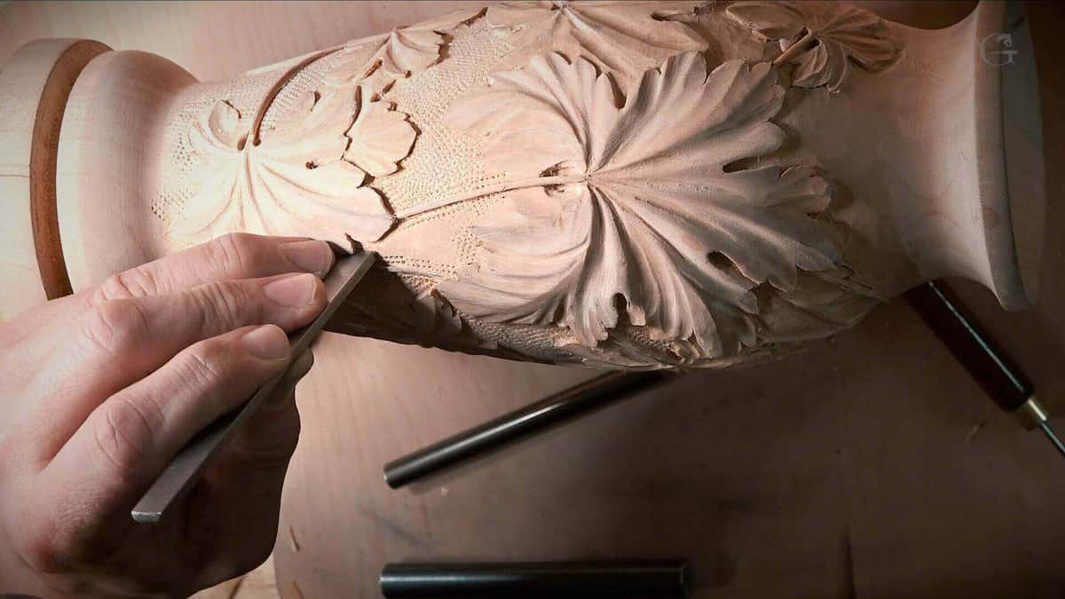 A flat chisel held almost level against a turned baluster carved with leaves, two gouges on the bench.