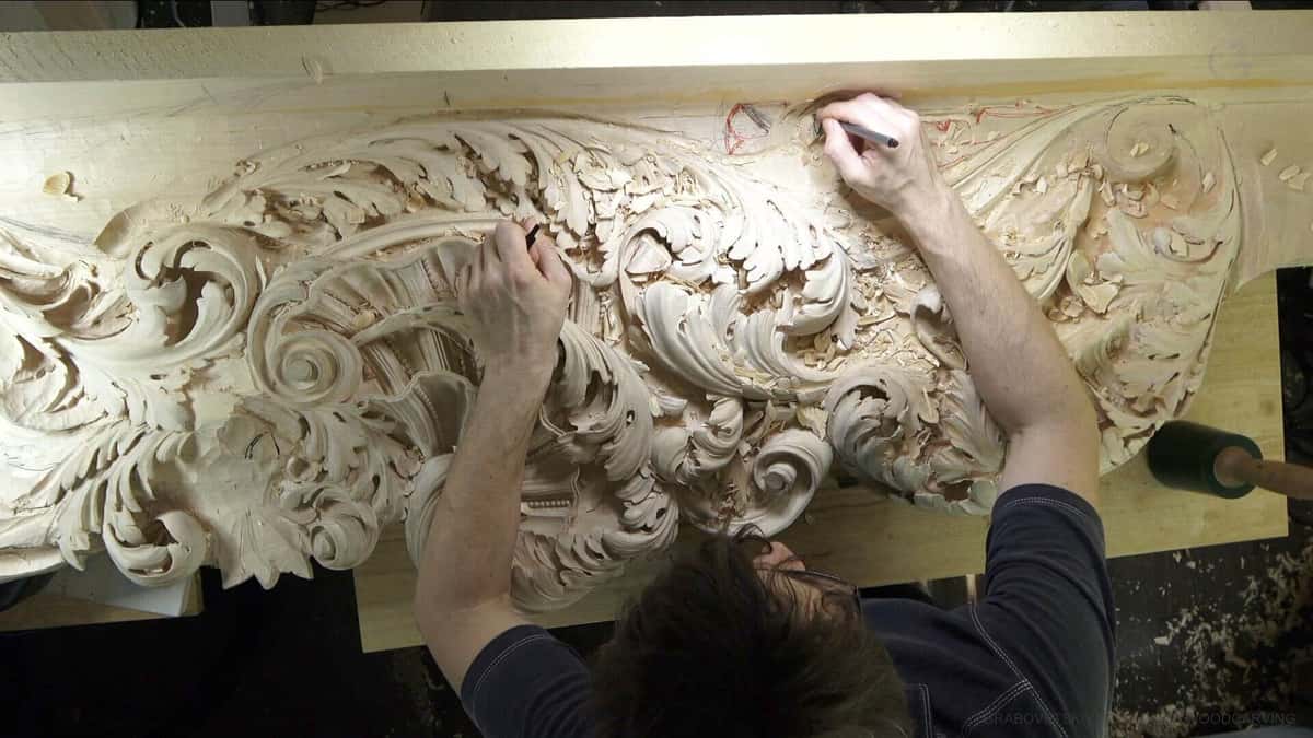 Overhead of a long acanthus frieze mid-carve, red pencil marks on the wood and chips everywhere.