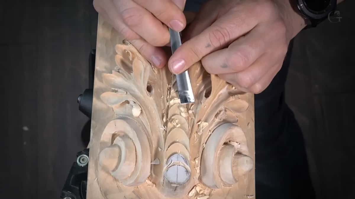 Hands press a flat chisel into a part-carved pale leaf panel gripped in a bench vise.