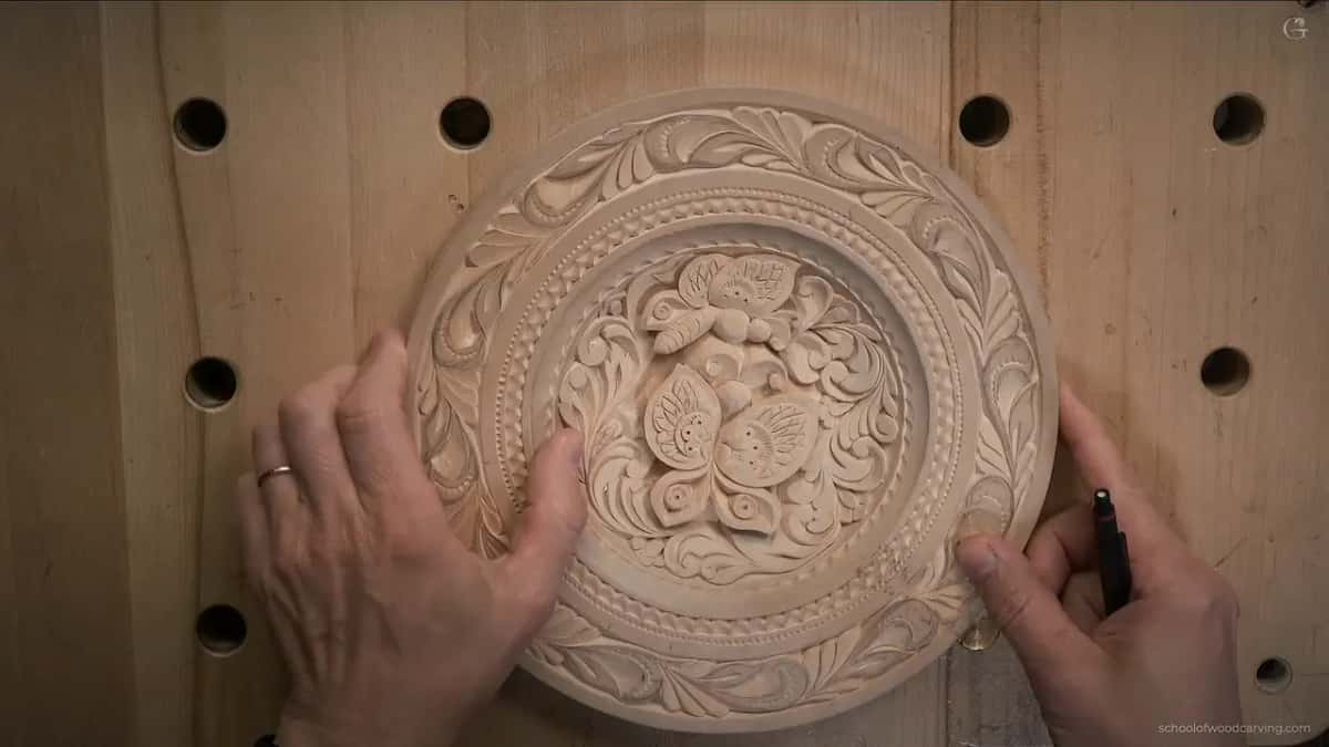 Hands hold a finished round plate carved in pale wood.