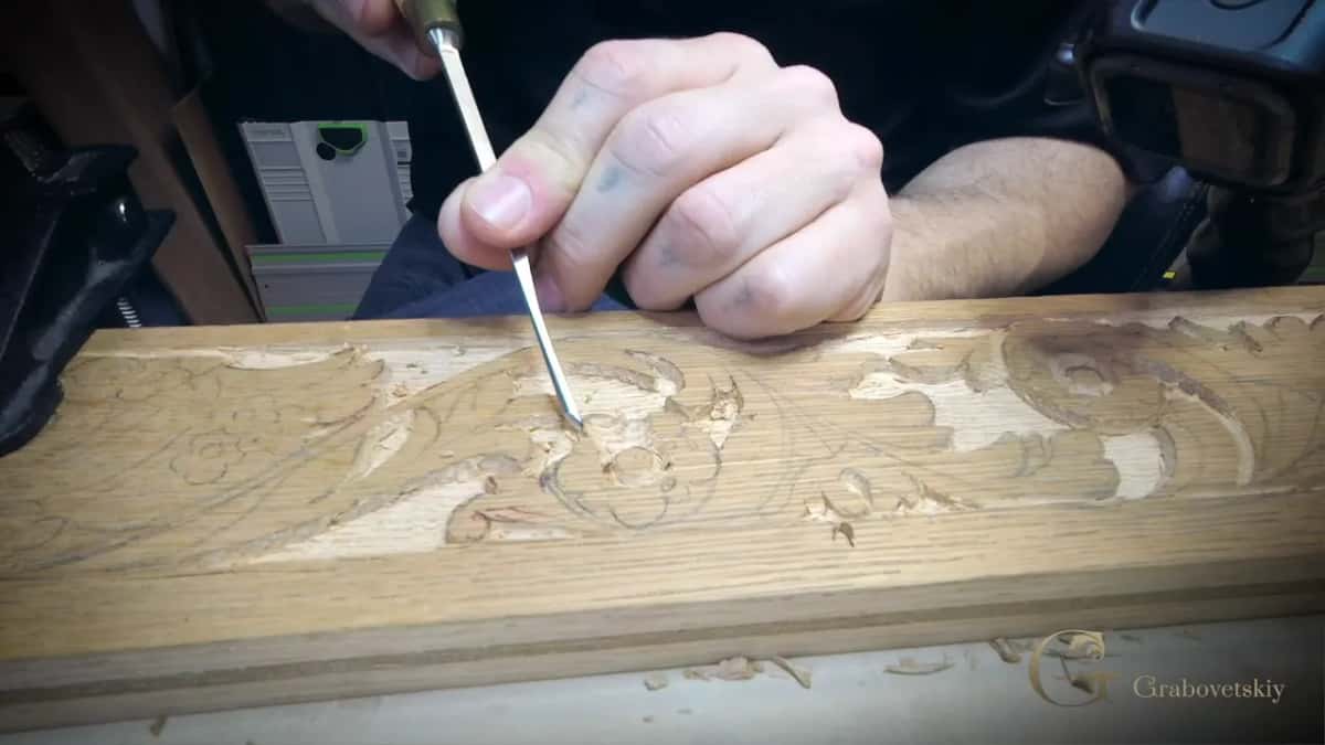 A hand drives a small gouge into a tan moulding strip, pencil scrolls still showing in the open grain.
