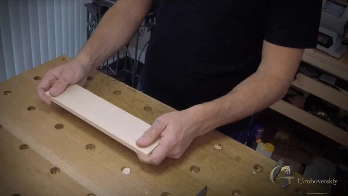 Hands set a narrow pale plank flat on an oak bench full of round holdfast holes.