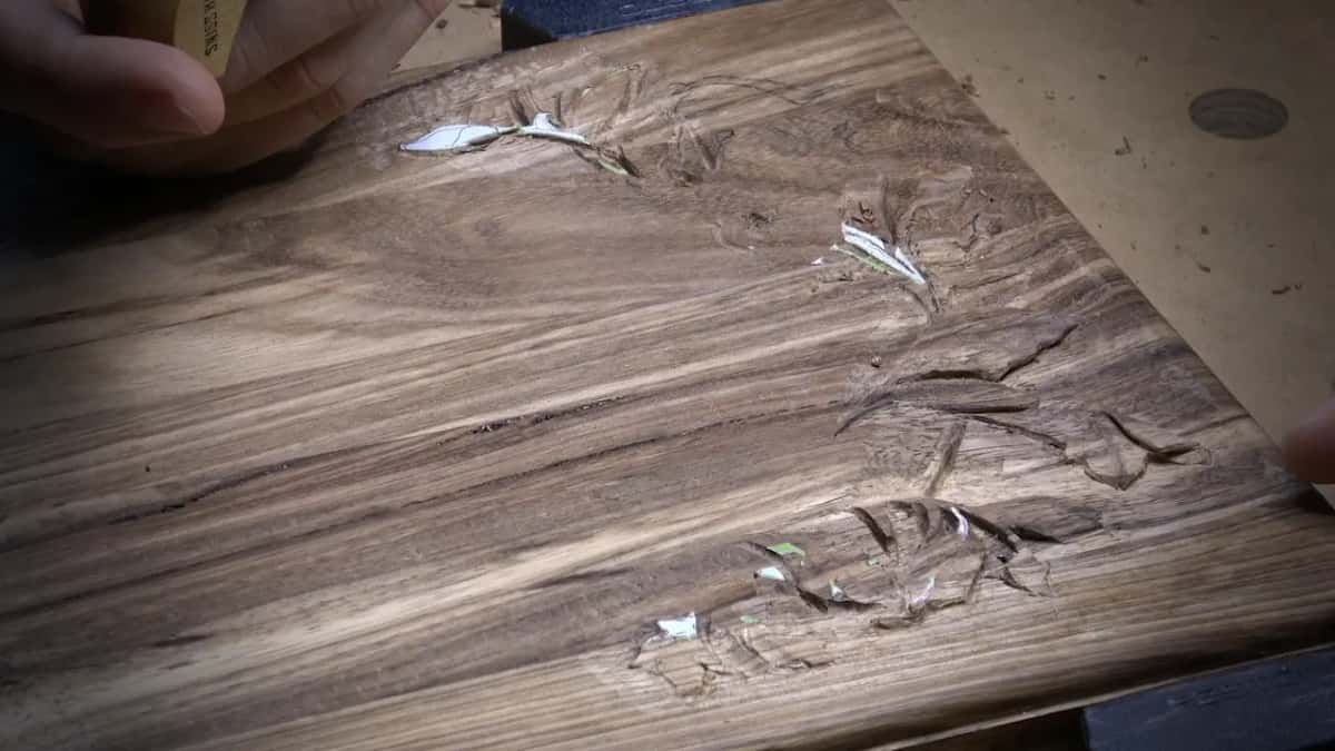 A brown board with wild streaked grain, leaf shapes roughed in and a mallet handle in the hand above.
