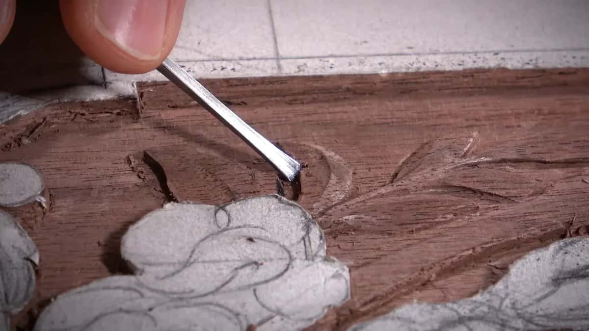 Close view of a flat chisel tip biting into a reddish-brown carved leaf, deep shadow at the point.