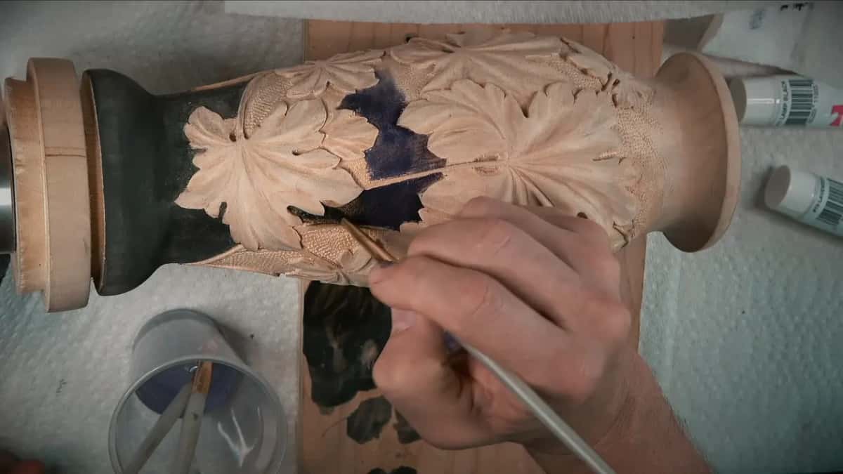 A brush lays dark pigment into the carved grape leaves of a turned vase, paint tubes beside it.