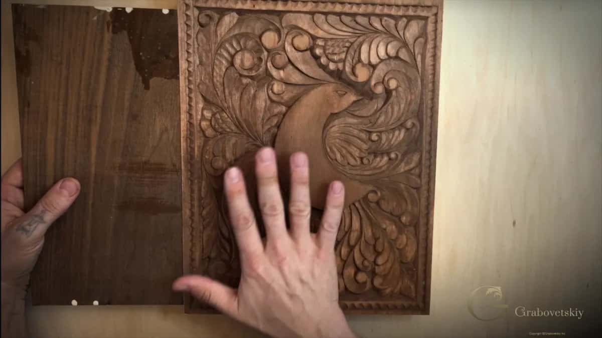 A hand rests on a finished dark-stained panel carved with a bird among acanthus, its plain back board alongside.