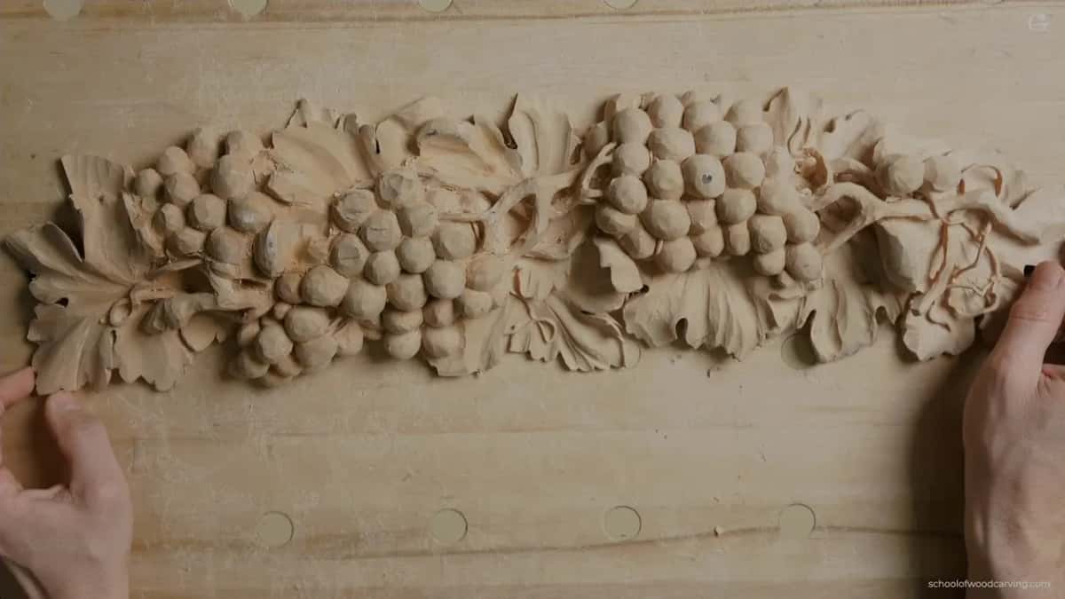 Hands hold the ends of a long grape onlay carved in pale wood, resting on the plywood bench.