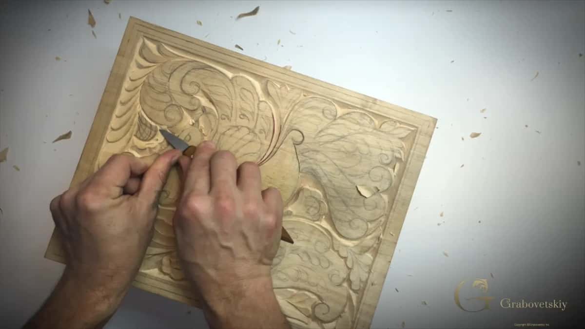 Two hands guide a small knife over a shallow acanthus panel carved into a pale curly board.