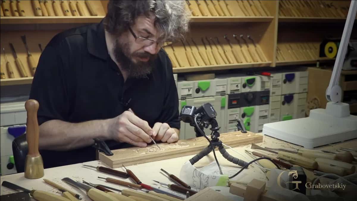 He bends over a long tan open-grained board with a fine tool, a brass-headed mallet standing among the gouges.
