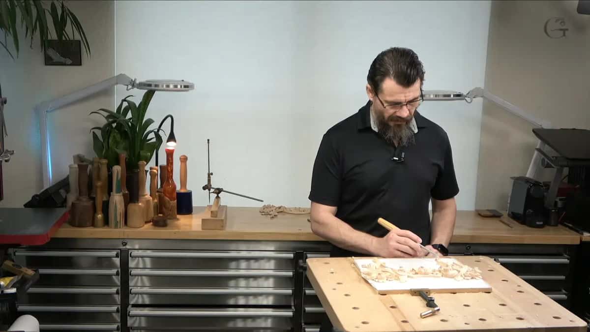 Wide studio view: he leans over a pale floral panel with a gouge, a row of turned mallets standing behind.