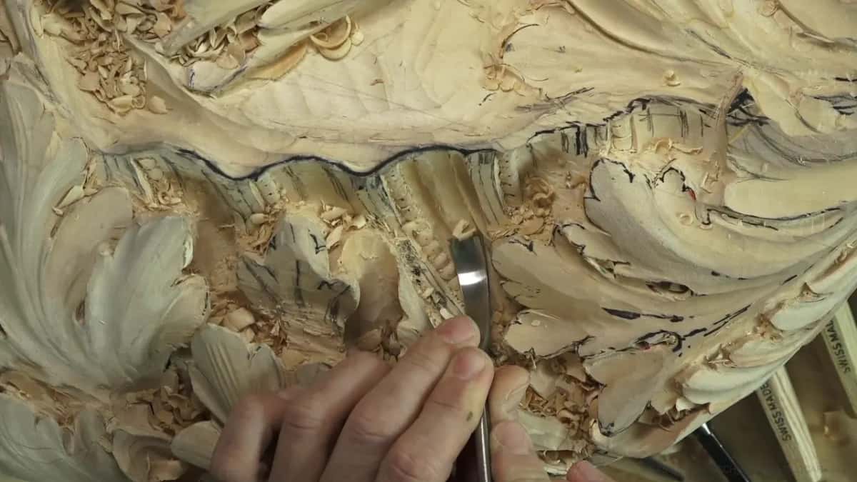 A gouge blade slicing a fluted channel between two carved leaves, chips piling in the hollow.
